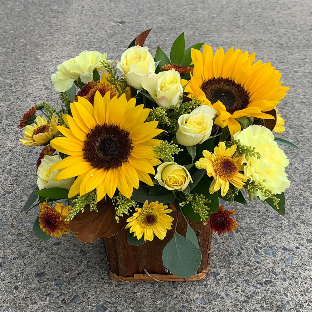 a yellow and brown floral arrangement in a brown wooden box. The arrangement is filled with sunflowers and yellow roses.